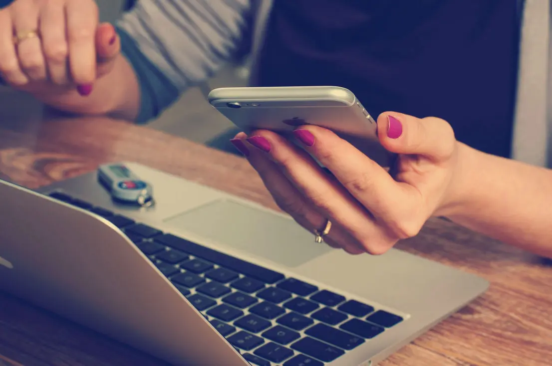 hand holding a smartphone, on a table stands a laptop